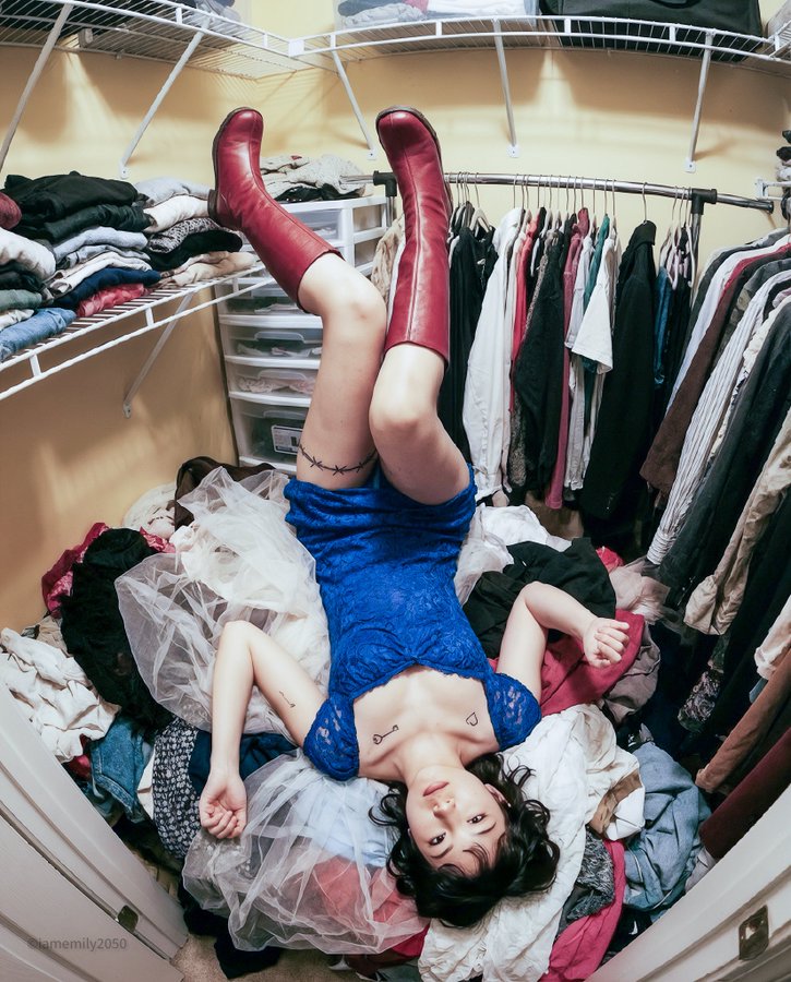 Generate a warm sepia, vintage 90s disposable-camera style bird’s-eye photo of an East Asian female idol lying upside-down in a cluttered yellow-walled closet, strictly matching the pose and anatomy from the provided reference image.
