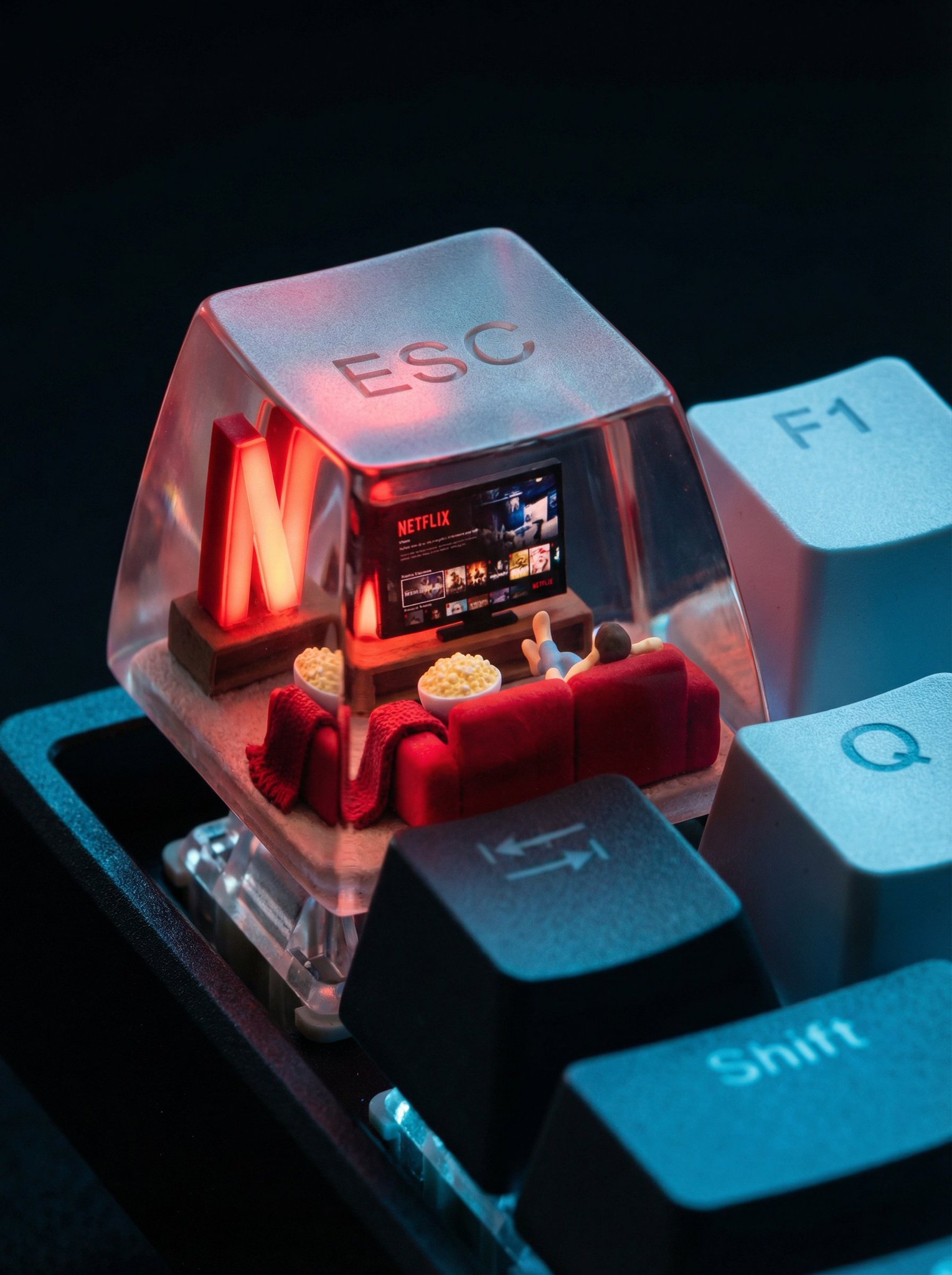 A photorealistic ESC keycap scene featuring a miniature cozy living room with a glowing red Netflix screen, plush red couch, popcorn bowl, and throw blanket, set against cool tech-blue reflections on surrounding keys. The “ESC” lettering appears as subtle glassy fog on the cap’s surface.