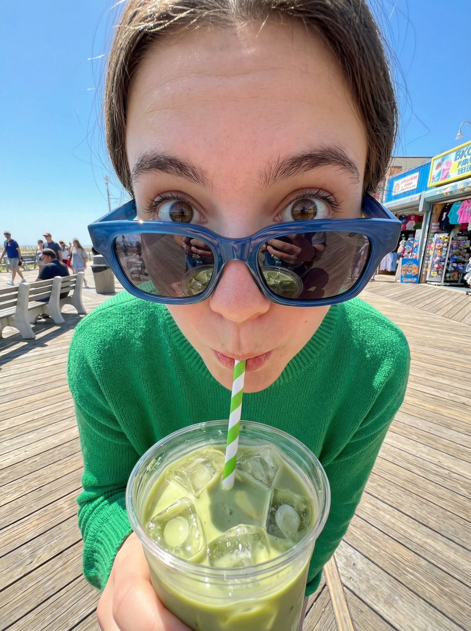 An extreme close-up, ultra-wide fisheye portrait of a young person sipping an iced matcha latte on a bright, sunny boardwalk with colorful stalls and passersby in the background.