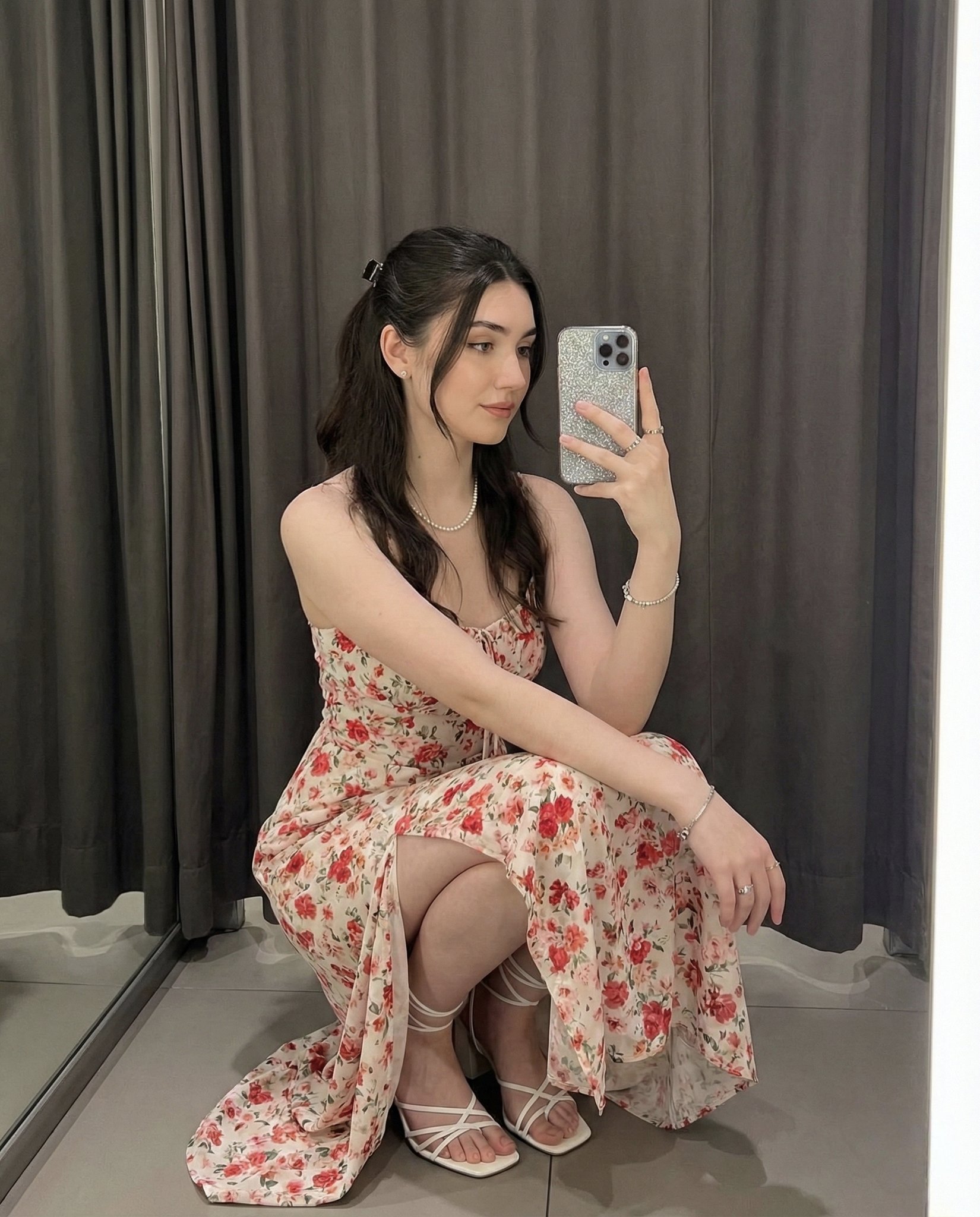 A young woman squats gracefully for a full-body mirror selfie in a softly lit indoor room with dark grey curtains and a reflective floor. Warm tones, medium-high clarity, and a glittery silver phone case complete the feminine, elegant social-media aesthetic.