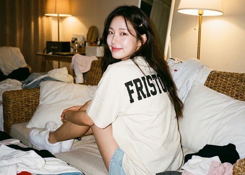 A hyperrealistic, warm vintage lifestyle interior photo featuring a girl seated on a woven sofa with her back turned, looking over her shoulder at the camera in a cozy, messy room.