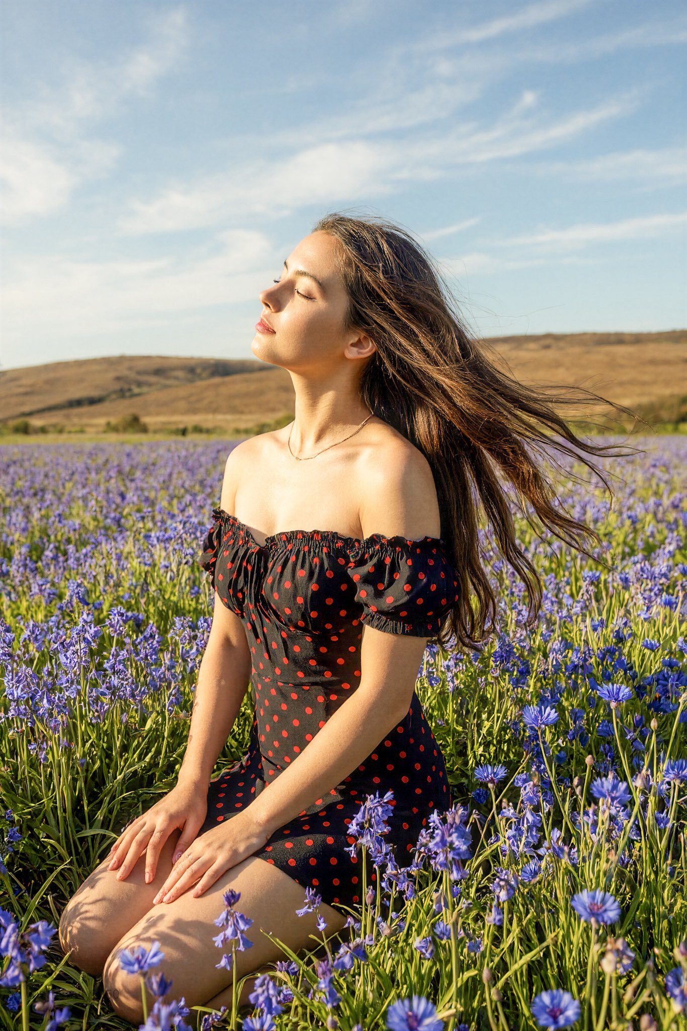 A serene portrait of a young woman kneeling in a dense blue wildflower field under hard, side-lit daylight, with windblown hair and distant rolling hills beneath wispy clouds.