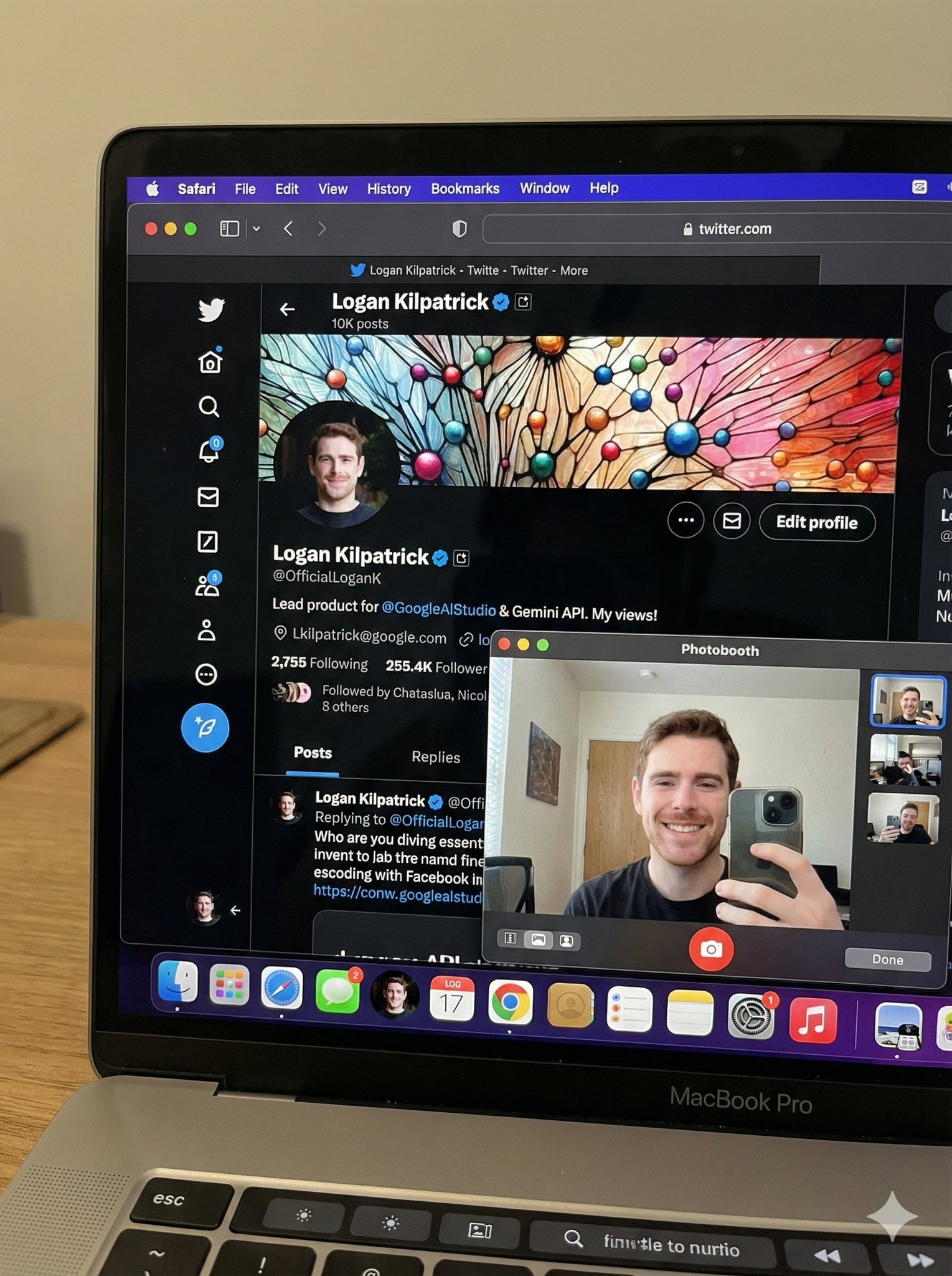A candid 3:4 phone photo of a MacBook Pro showing X open in Safari alongside Photo Booth capturing a webcam selfie. The scene includes the correct account UI details, visible profile picture in the dock/sidebar, and readable text with no gibberish.