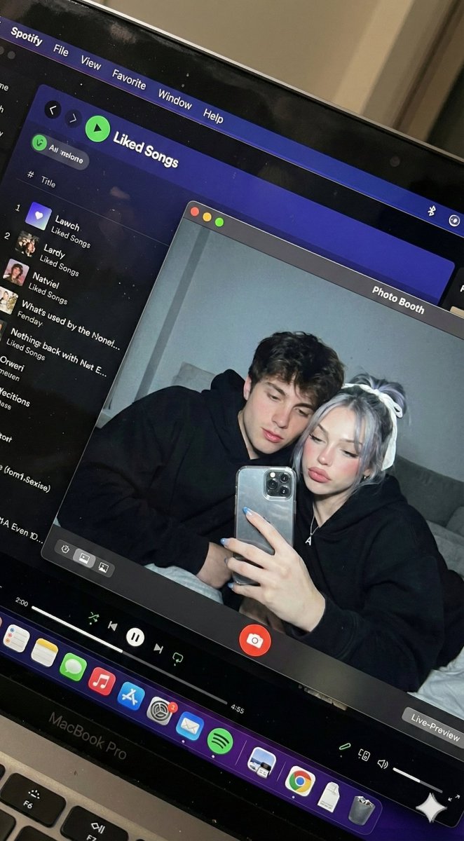 Create a hyper-realistic downward-angled photo of a MacBook screen showing a Photo Booth live preview of a couple in a dim bedroom, with authentic screen reflections, dust, moiré, and fingerprints.