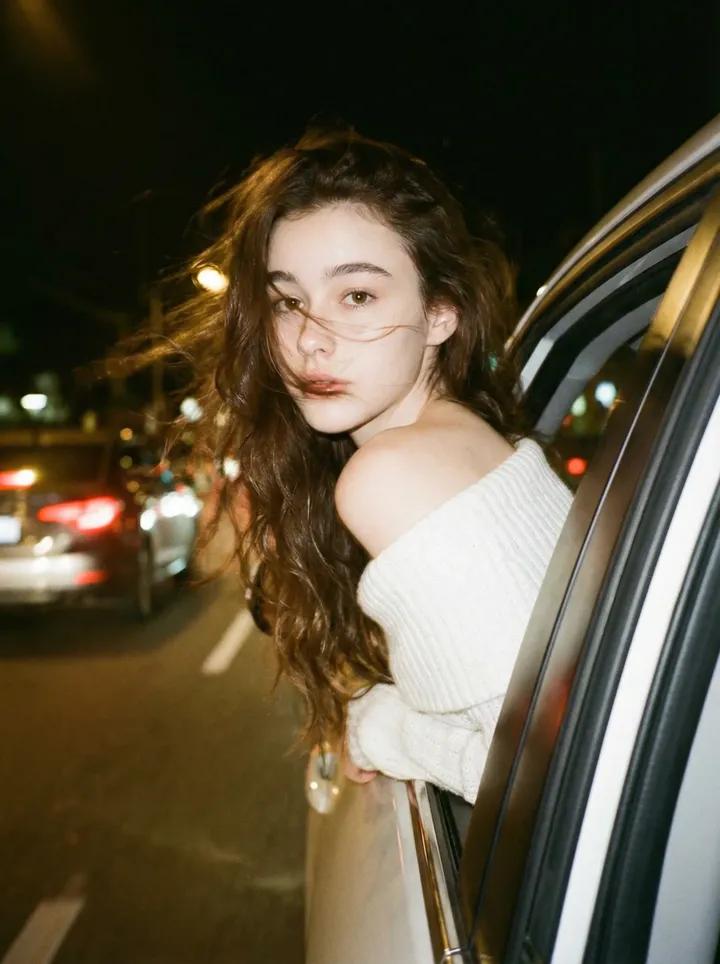 Create a dark, dreamy, blurry vintage-style night photo of you leaning your back out of a car window, with a windy atmosphere. The look features brown long wavy hair and an off-shoulder white knitted top while keeping facial details and head positioning unchanged.