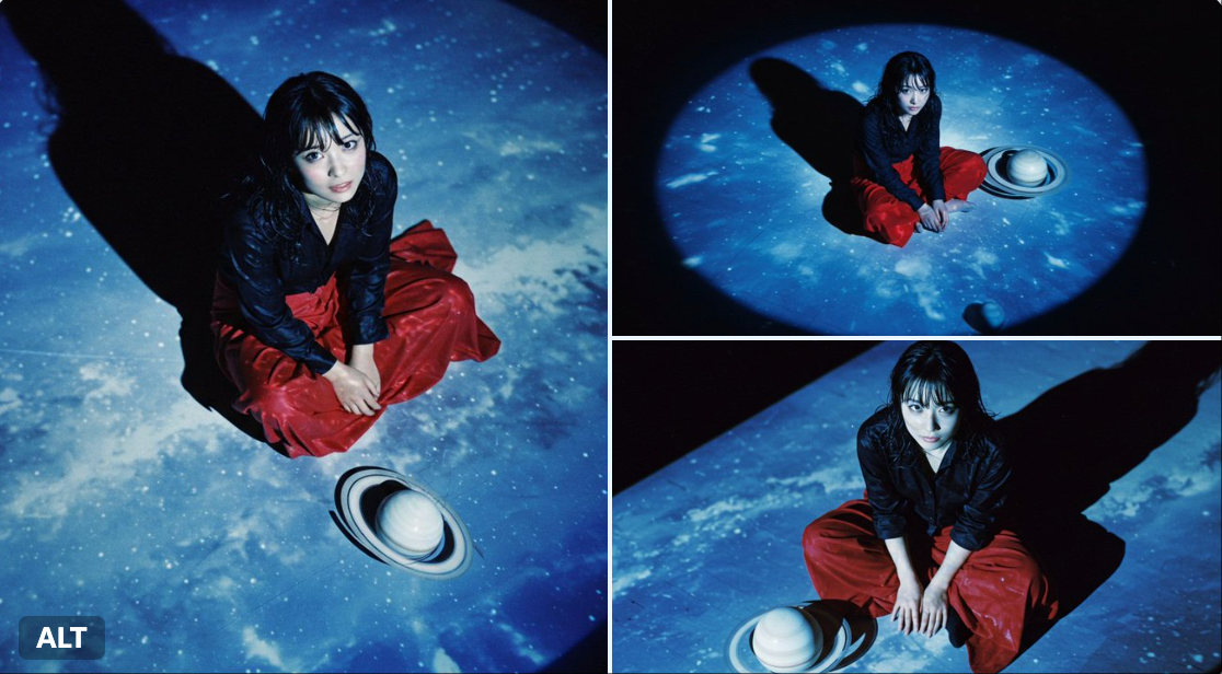A cinematic overhead close-up shot with bluish tones and realistic ISO100 film shadows, featuring a Japanese film actress seated on a black floor under a projected blue sky. The scene includes striking light and shadow, a long red skirt, and a white Saturn model for a surreal movie-still feel.